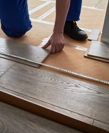 Close up of man construction worker checking distance from wall to laminate board. Male builder using tape measure while installing laminate flooring in apartment under renovation.